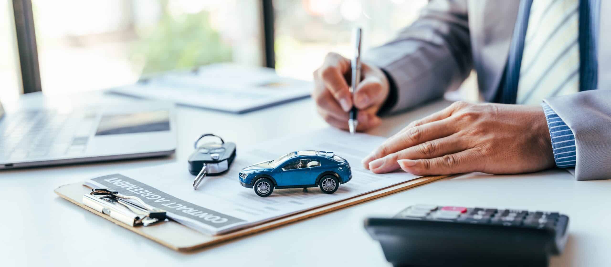 A person in a suit is signing an auto insurance document on a clipboard with a small toy car, a calculator, car keys, and a laptop on the desk.