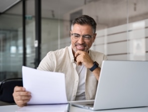 A man wearing glasses and a beige shirt is sitting at a desk, smiling while looking at papers in his hand. At home, with a laptop open beside him, he seems content amid familiar surroundings.
