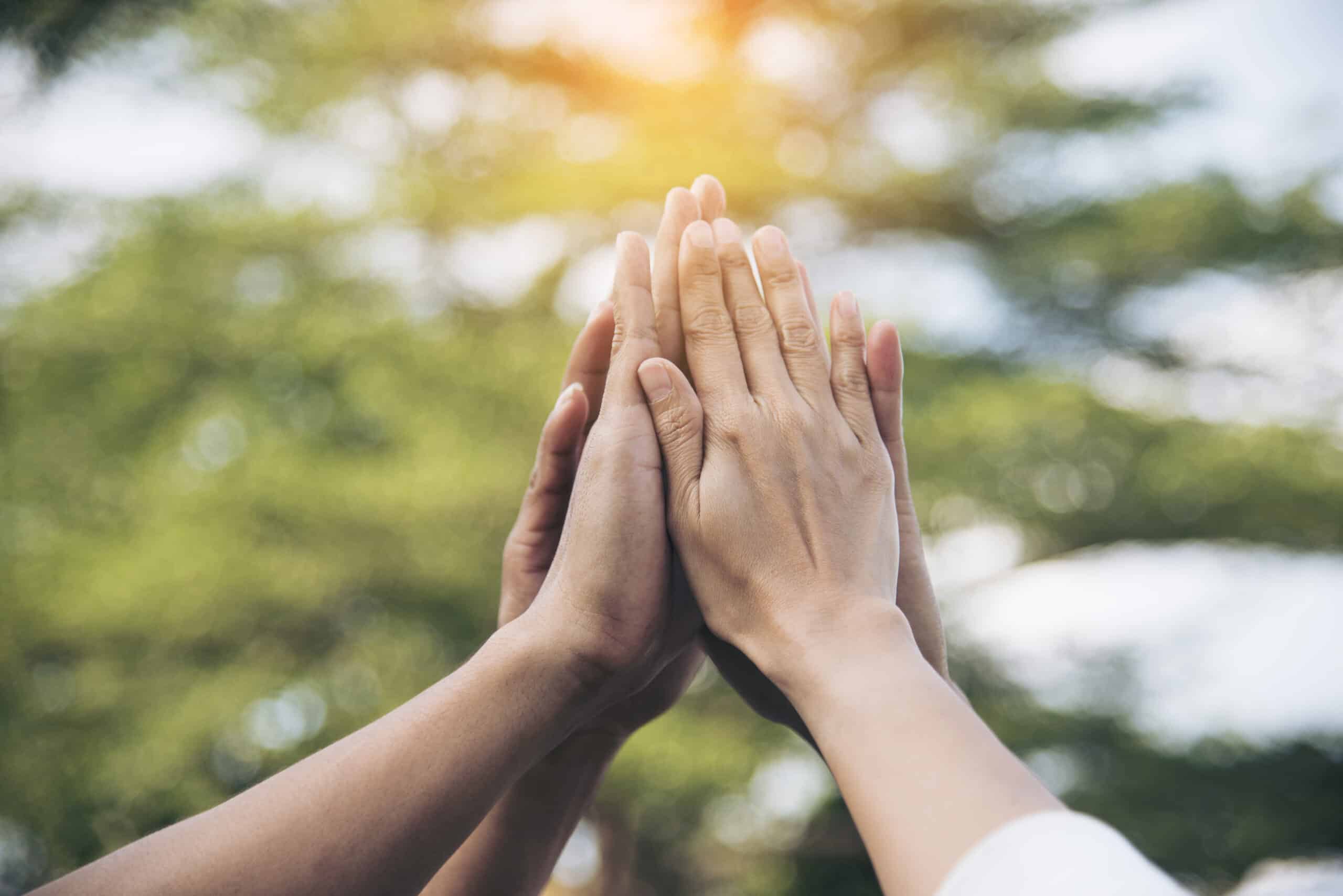 Four hands touching each other with fingers pointing upward, symbolizing the impact of unity, set against a blurred background of trees and sunlight.