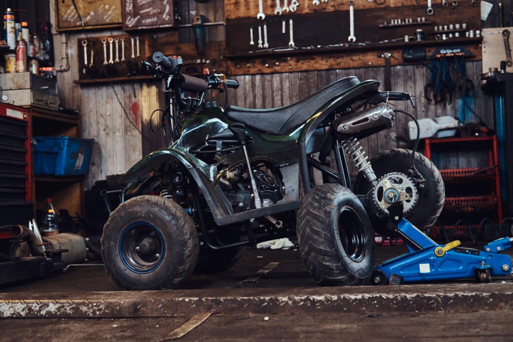 A four-wheeled all-terrain vehicle is elevated on a blue jack in a workshop, with tools hanging on the back wall.