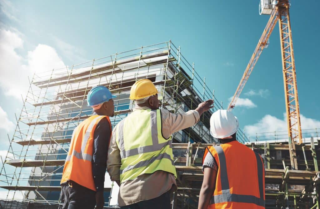 Three construction workers wearing helmets and safety vests converse about Builder's Risk Insurance while looking at a building under construction, with scaffolding and a crane in the background.