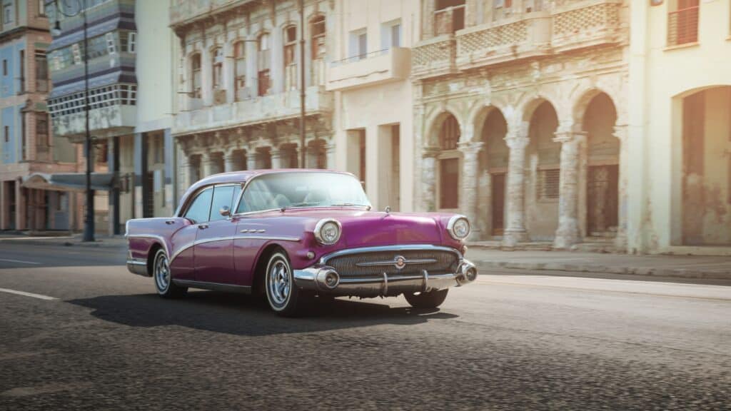 A vintage purple car insured by Classic Auto Insurance drives along a street lined with old, weathered buildings on a sunny day.