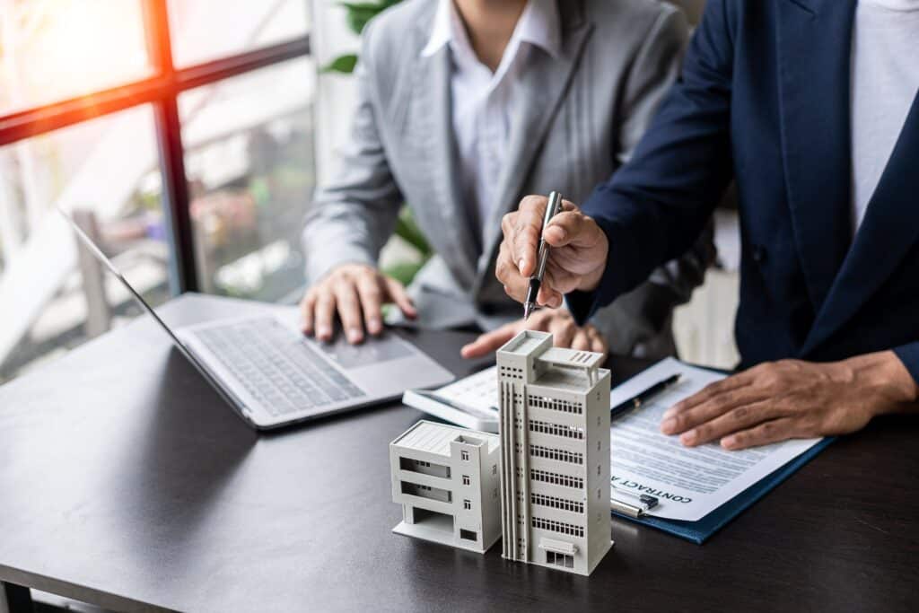 Two people in business attire sit at a desk with a laptop, architectural model, and contract. One points to the model of their commercial property while the other writes on the insurance contract.