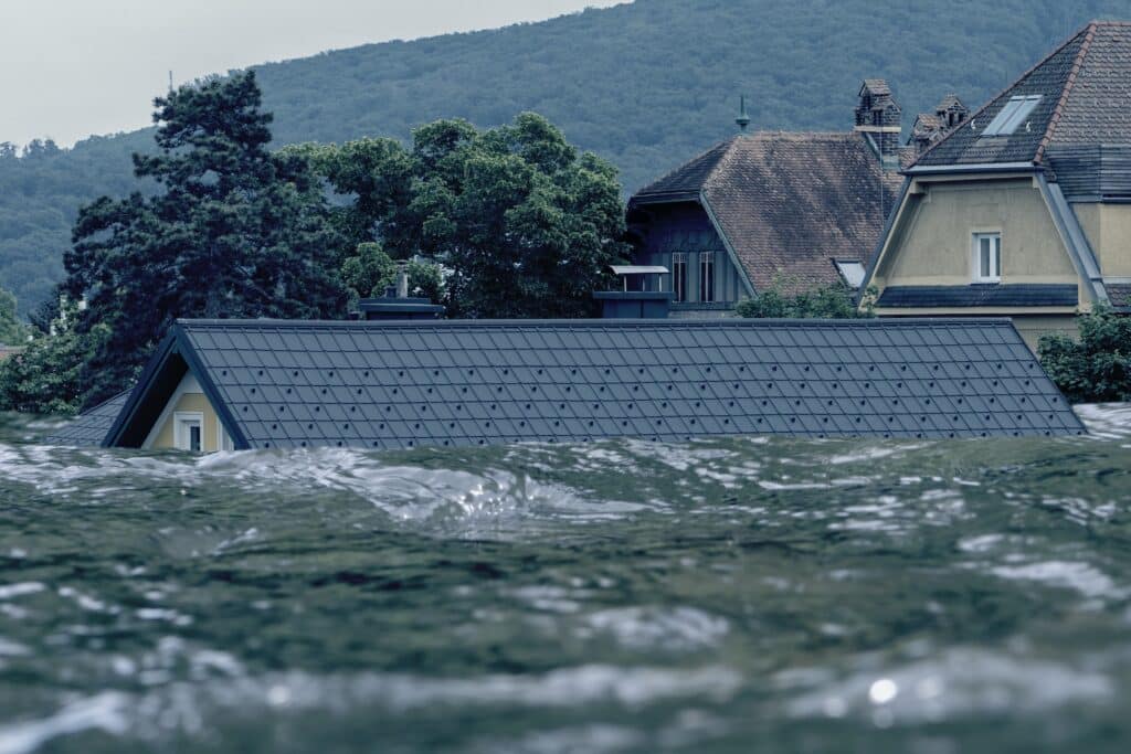 Houses partially submerged in floodwaters, with only rooftops visible under a cloudy sky and hilly background—a stark reminder of the need for comprehensive flood insurance.