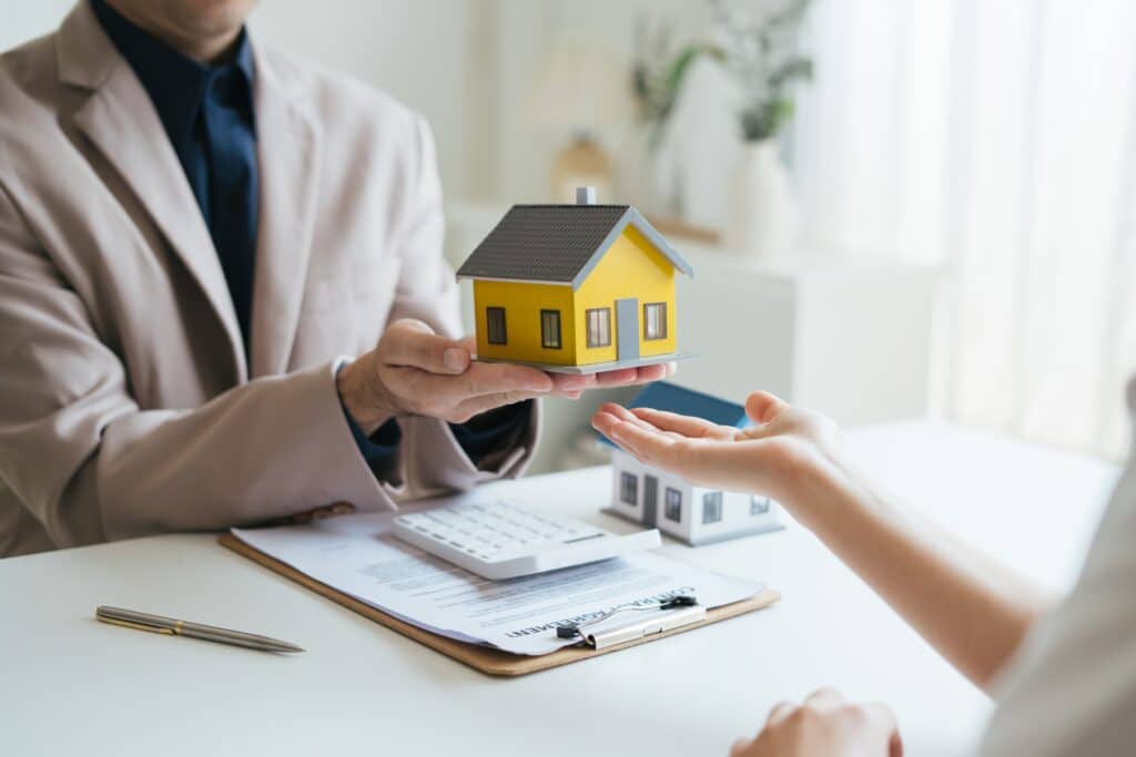 A person in a suit hands over a model yellow house to another individual, with documents and a white calculator on the table in the background, possibly discussing General Liability Insurance.