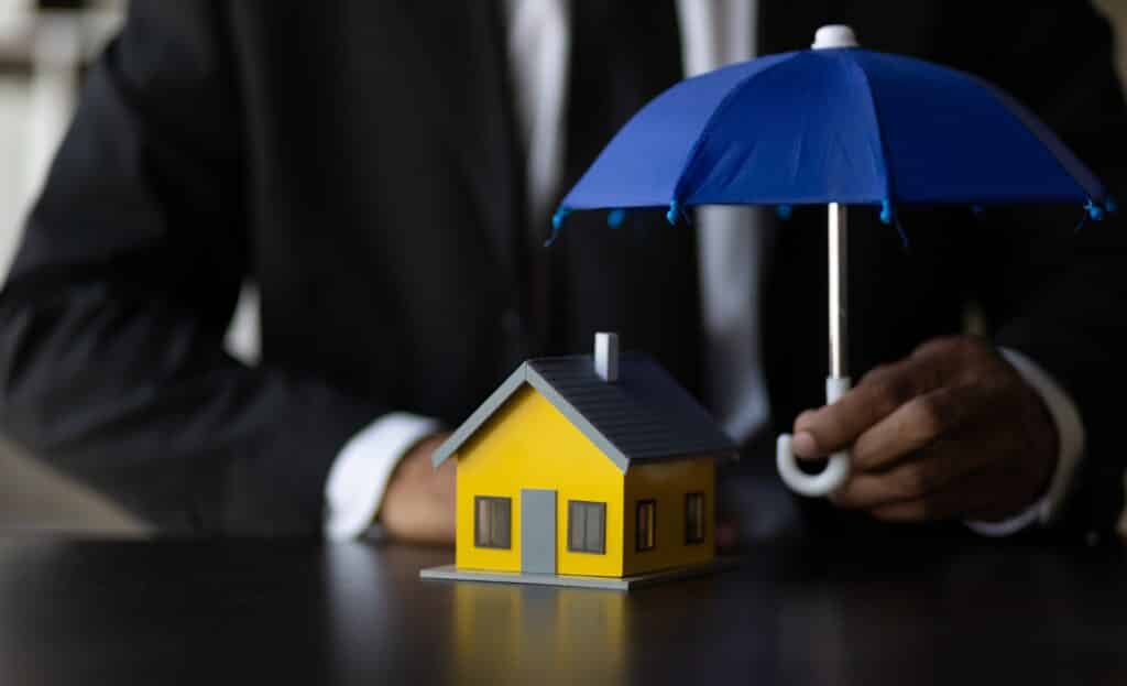 A person in a suit holds a blue umbrella over a small model yellow house on a table, symbolizing the protection offered by homeowners insurance.