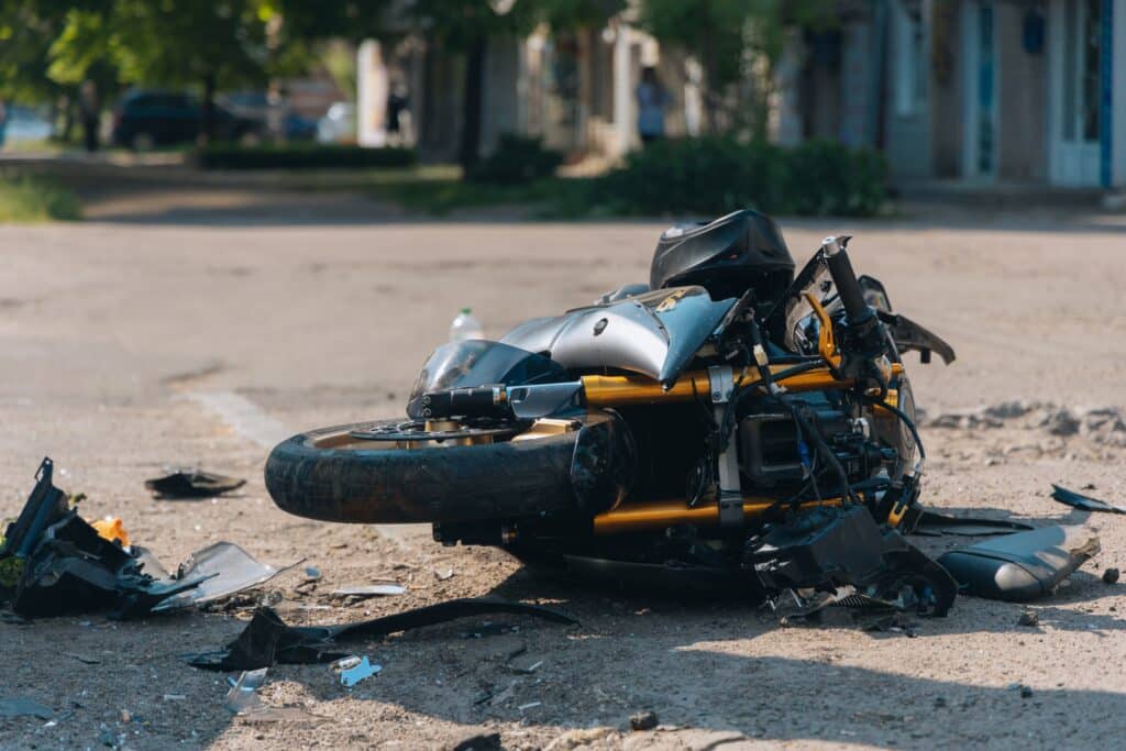 A damaged motorcycle lies on its side amid scattered debris, painting a grim accident scene. Trees and buildings stand in the background, underscoring the urgency for comprehensive motorcycle insurance.