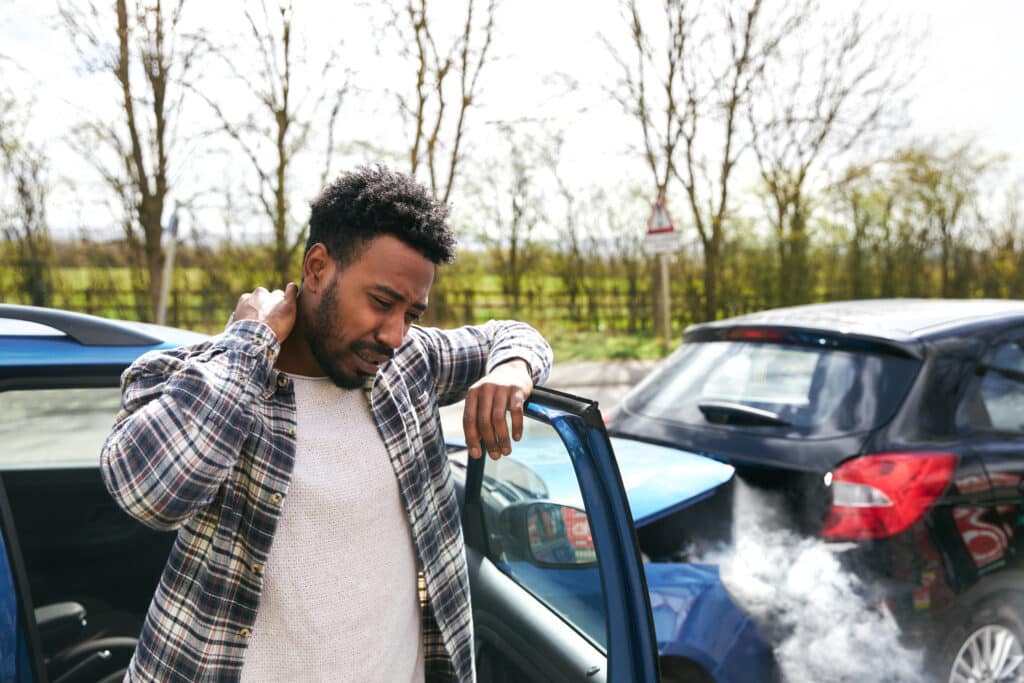 A man stands by the open door of a blue car, holding his neck, in front of a car accident scene with smoke coming from the vehicles. The rear-end collision on a rural road serves as a stark reminder of the importance of having reliable auto insurance.