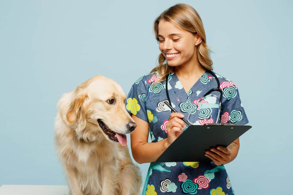 A smiling veterinarian in a floral scrub top holds a clipboard and stands next to a golden retriever in a light blue room, ready to discuss pet health coverage.