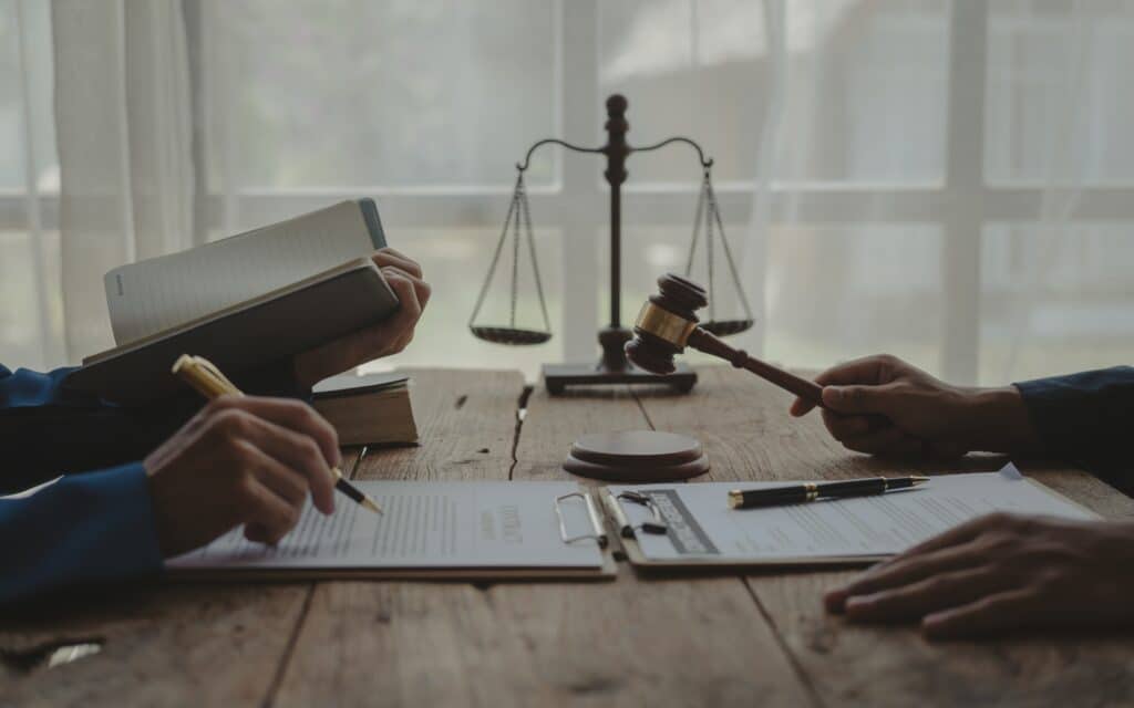 Two people work at a wooden table with documents, a gavel, and scales of justice. One holds a pen, the other a book, discussing professional liability under natural light streaming through the window.