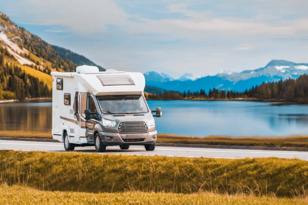 A motorhome is parked on a scenic road with a lake and mountains in the background under a clear sky, showcasing the peace of mind that comes with comprehensive RV insurance.