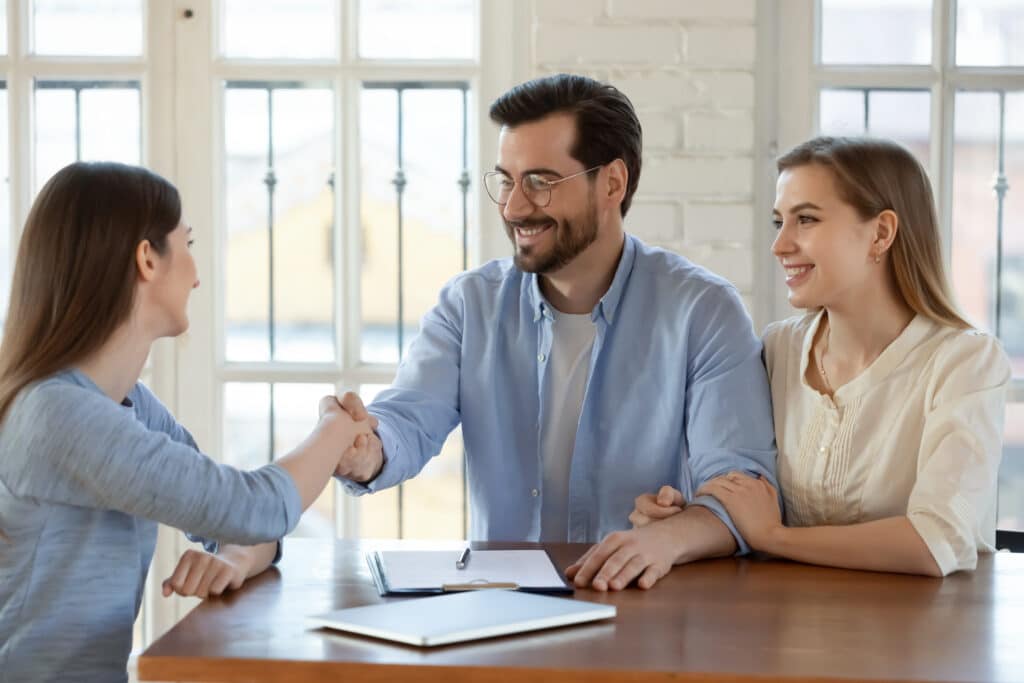 Two women and a man sit at a table in a bright room. The woman on the left and the man shake hands, while the woman on the right smiles and touches the man's arm. Papers about Renters Insurance and a pen are on the table.