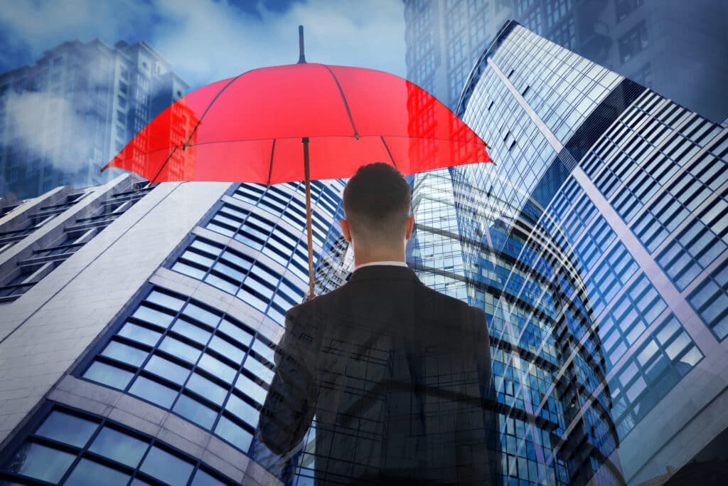 A person holding a red umbrella stands in front of multiple high-rise buildings, contemplating their vastness under a cloudy sky, representing a umbrella liability insurance concept.