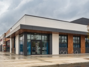 Modern commercial building with large glass windows and a flat roof on a cloudy day. The structure, which combines elements often seen in a contemporary home, features white, brown, and dark gray exterior panels. The area is wet from recent rain.