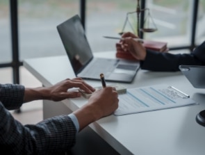 Two people are sitting at a desk in a cozy home office. One is signing a document while the other looks on. A laptop, a clipboard with a form, and a pair of scales are neatly arranged on the desk.