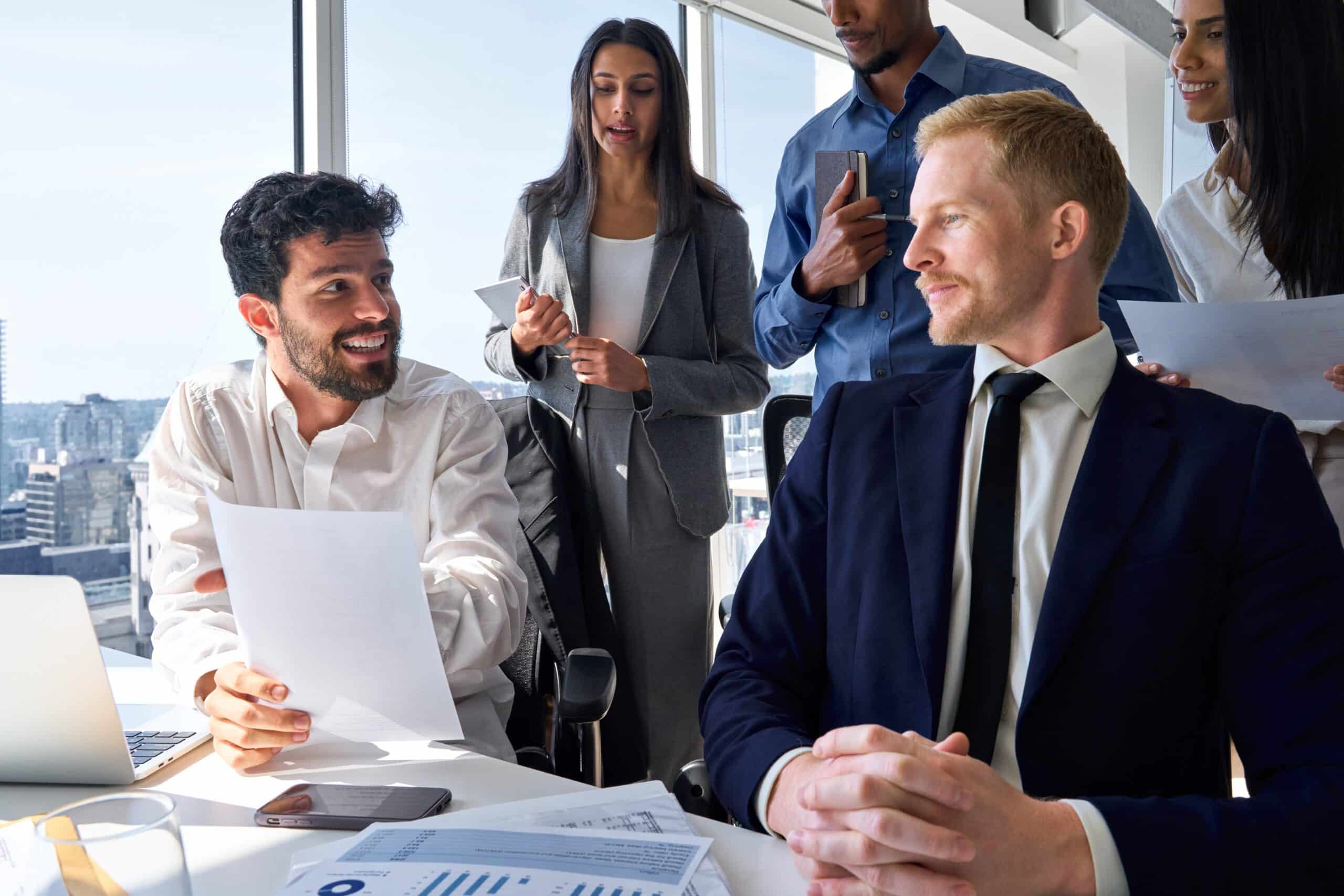 Five business professionals in a meeting room: two seated men are deep in discussion about small business insurance, while three standing colleagues observe and hold documents, focused on protecting your business from potential insurance gaps.