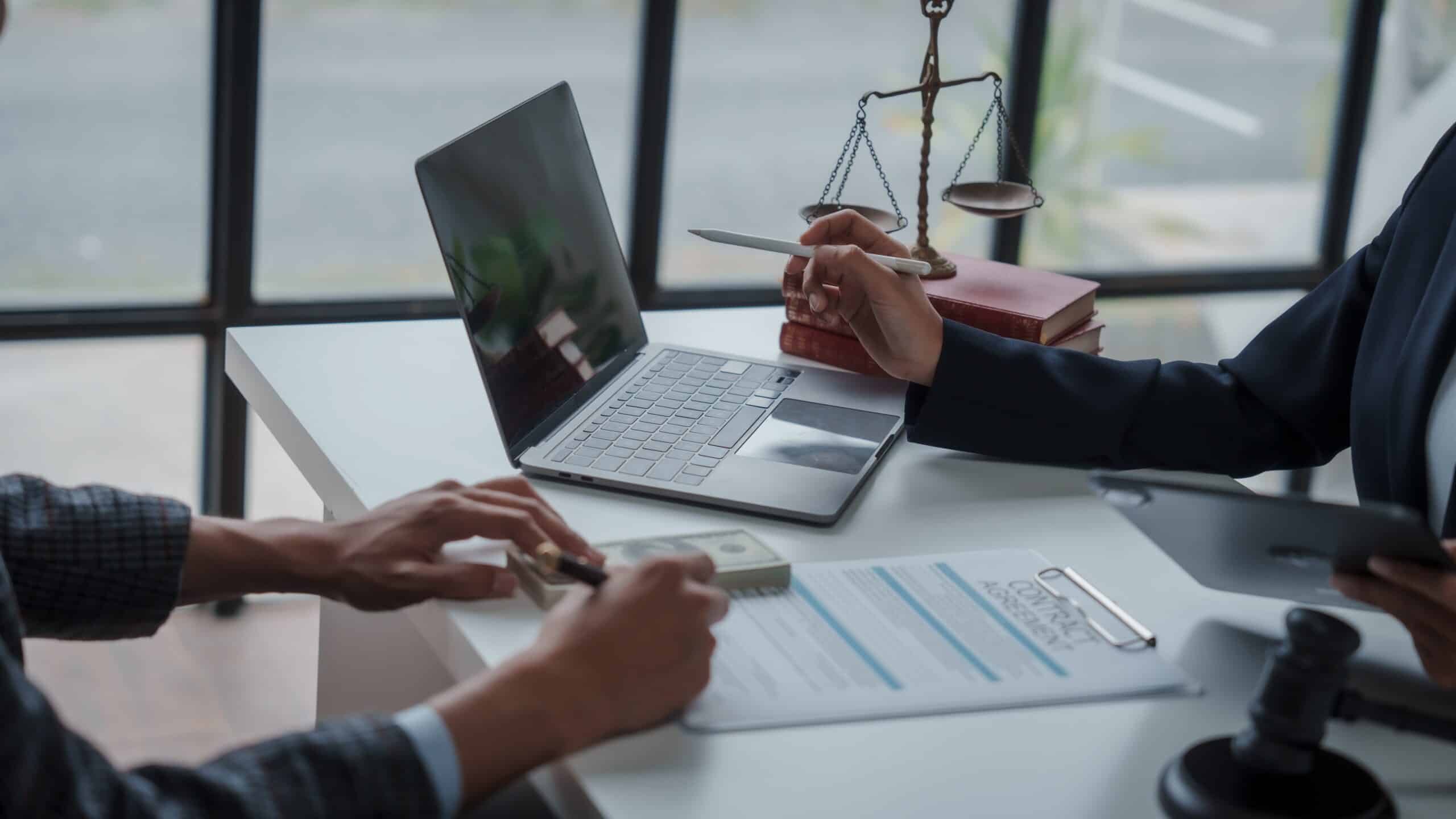 Two people discussing legal documents at a desk with a laptop, scales of justice, and a clipboard. One person is holding a pen and the other is reaching for a stack of cash, possibly finalizing an insurance policy that includes liability coverage.