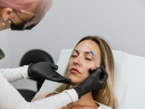 A person with black gloves marks another person's forehead with a blue pen as they lie on a medical bed, preparing for a cosmetic procedure that feels almost as comfortable as being at home.