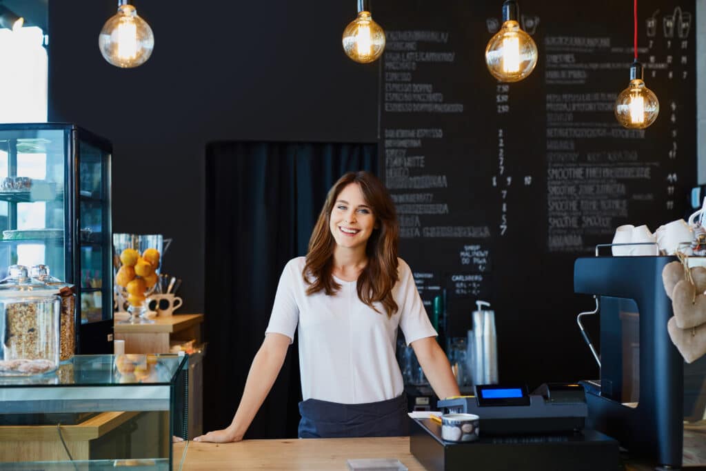 A woman stands behind the counter in a café, smiling, with hanging lights and a menu board in the background. She is wearing a white shirt and an apron, embodying the essence of small business insurance success.