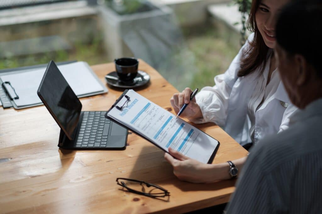 Two people at a wooden table discussing a document on a clipboard, likely related to large business insurance. A laptop, glasses, and a cup of coffee are also on the table.