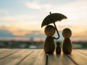 Wooden figures of a family holding an umbrella stand on a wooden surface against a blurred sunset background, evoking the warmth and security of home.
