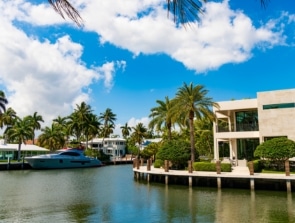 A modern waterside home with large windows, surrounded by palm trees under a blue sky with scattered clouds. A boat is docked nearby on the calm water.
