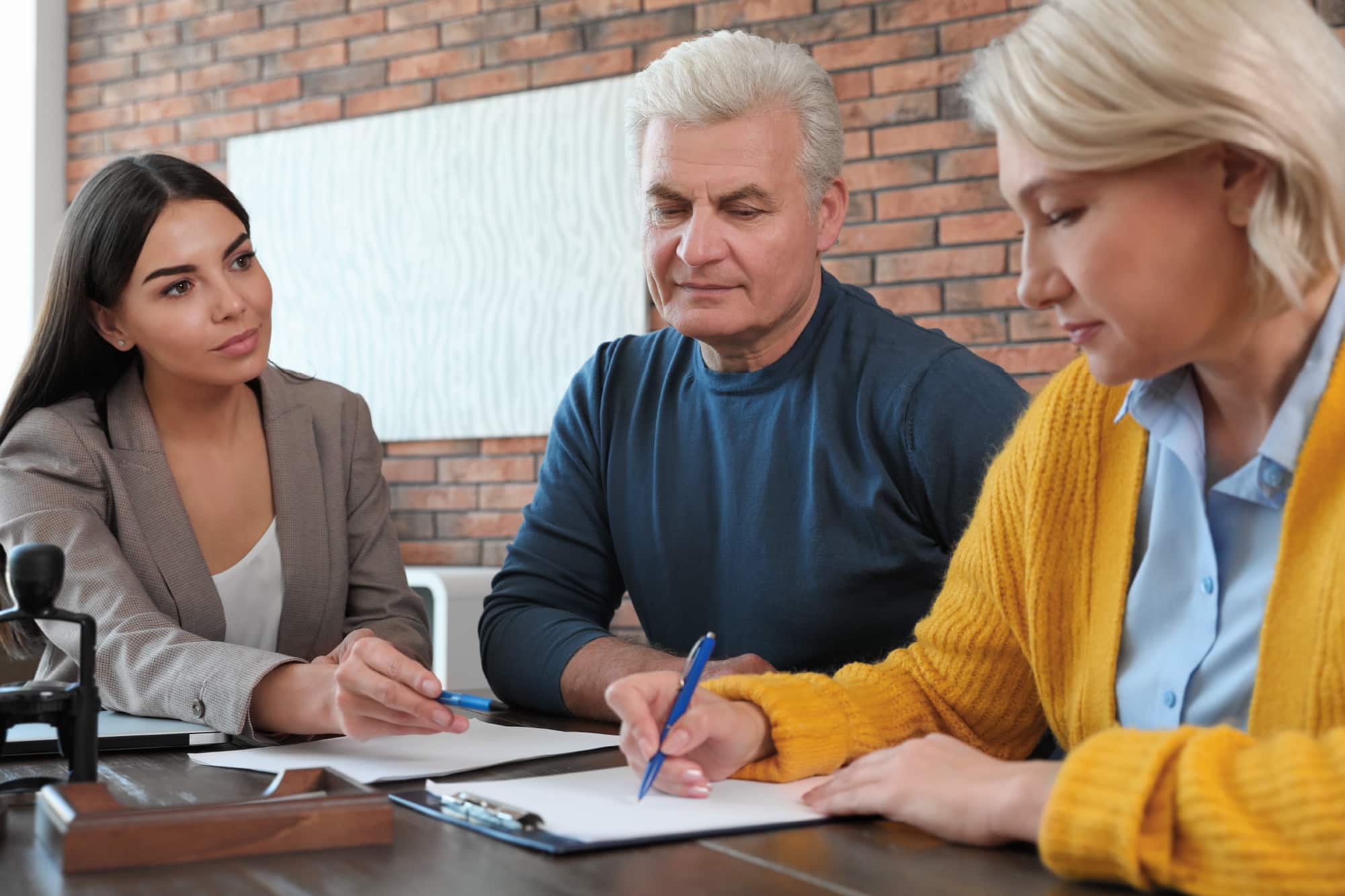 Three public adjusters sit at a table discussing documents, with one holding a pen, in a room with brick walls.