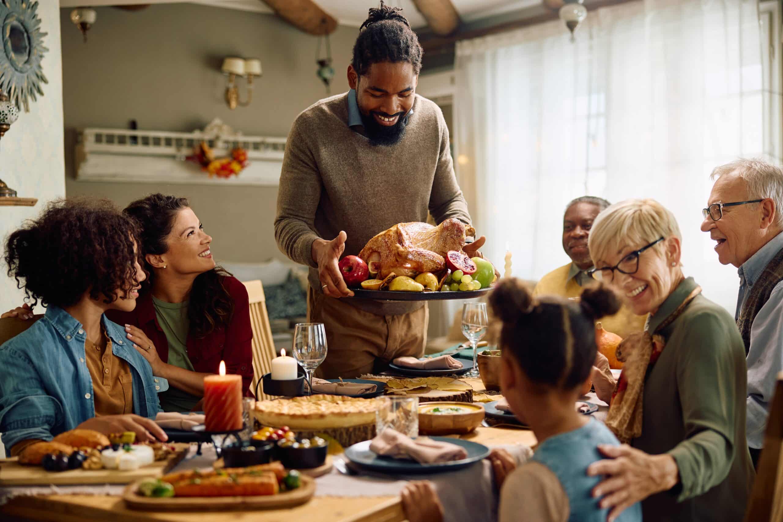 A man serves a roasted turkey to a group of smiling family members gathered around the dinner table, capturing the warmth and joy of Thanksgiving.