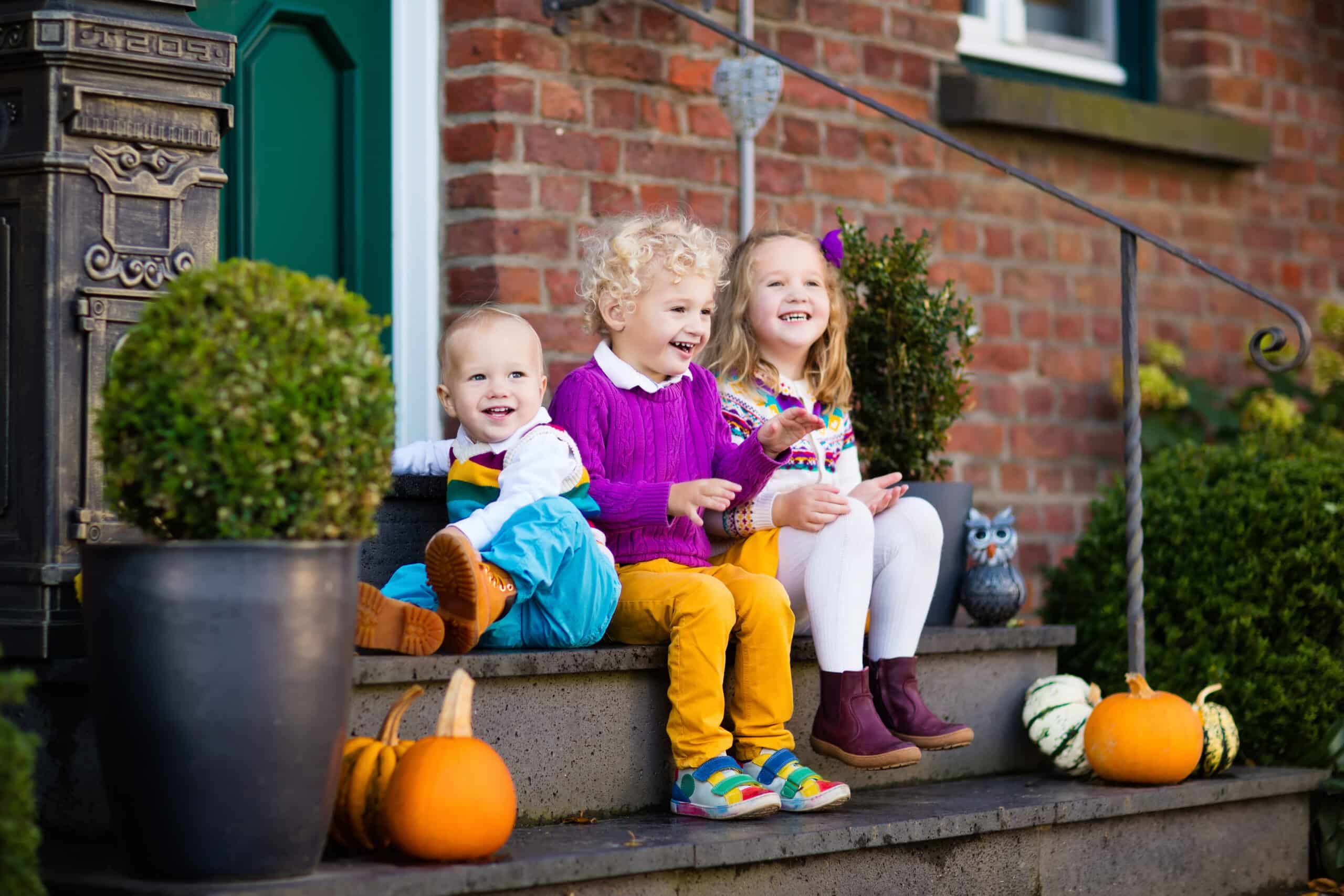 Three children sit on porch steps beside small pumpkins and greenery, smiling warmly. A brick wall and a green door, hinting at holiday safety, create a cozy backdrop for this cherished moment.