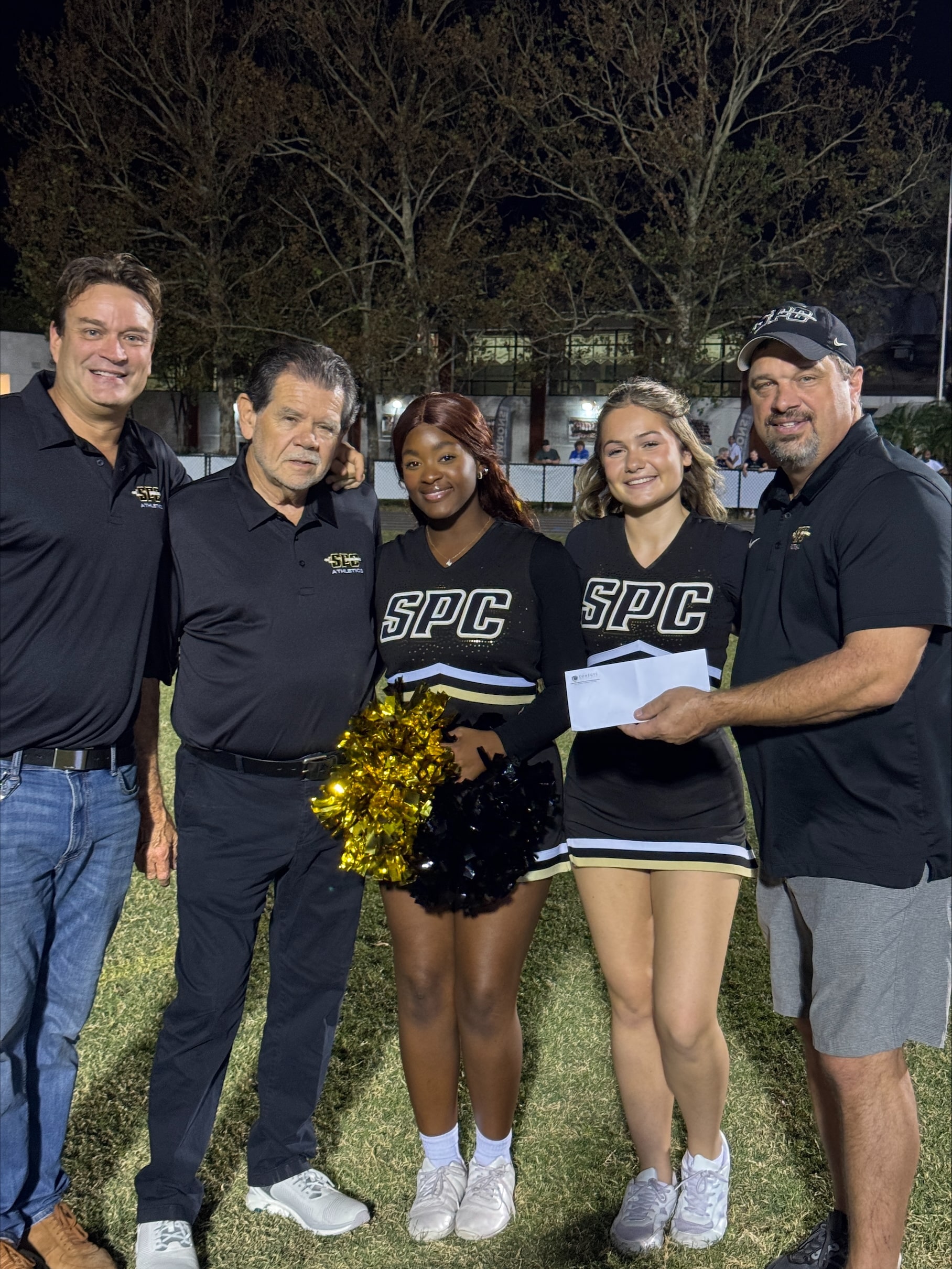Five people, including two cheerleaders with pom-poms, stand outdoors. Three men in black shirts, one holding an envelope marked 'donation,' smile warmly at the camera. Against a backdrop of trees, their gathering celebrates community support for St. Petersburg Catholic High School.