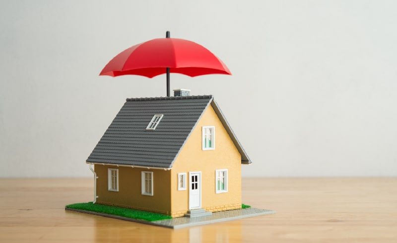 A small house model with a red umbrella suspended above it on a wooden surface.
