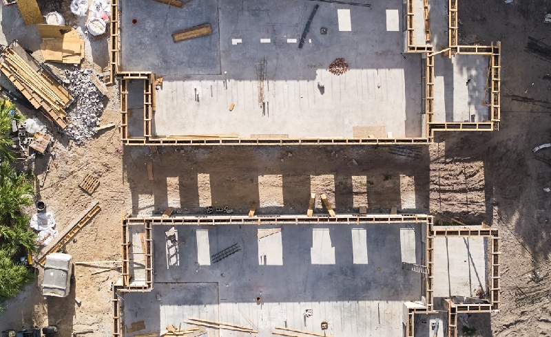 Aerial view of a construction site with concrete foundations and wooden frames set for two buildings. Equipment and materials are scattered around the area.