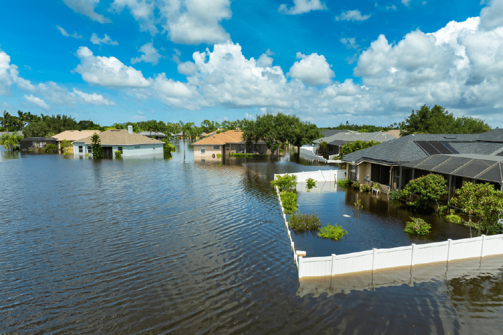 A flooded residential area with water covering lawns and reaching houses. Several trees and fences are partially submerged. The sky is cloudy with patches of blue.