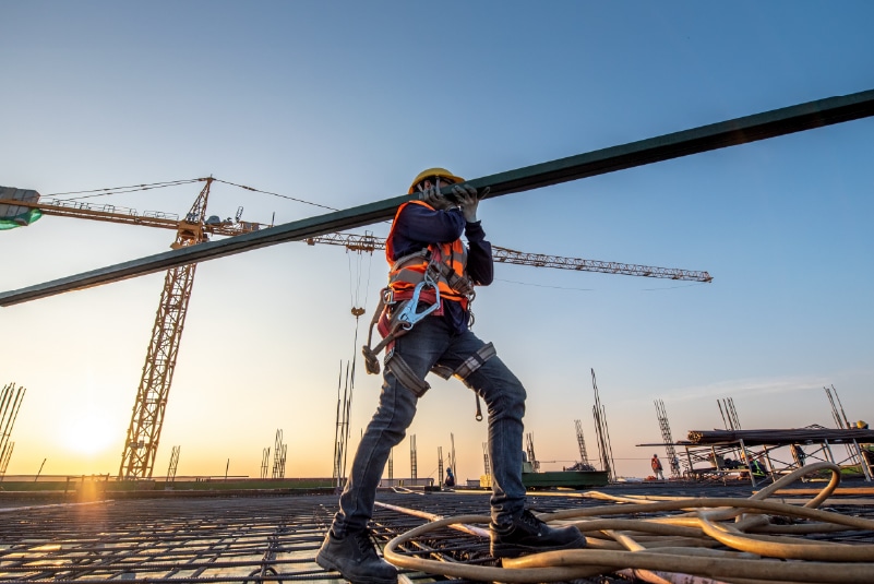 At sunrise, a construction worker in a safety vest and helmet expertly shoulders a metal beam on the bustling building site, setting the foundation for industry progress against a crane-studded skyline—a scene where every detail matters, just like crafting the perfect landing page.