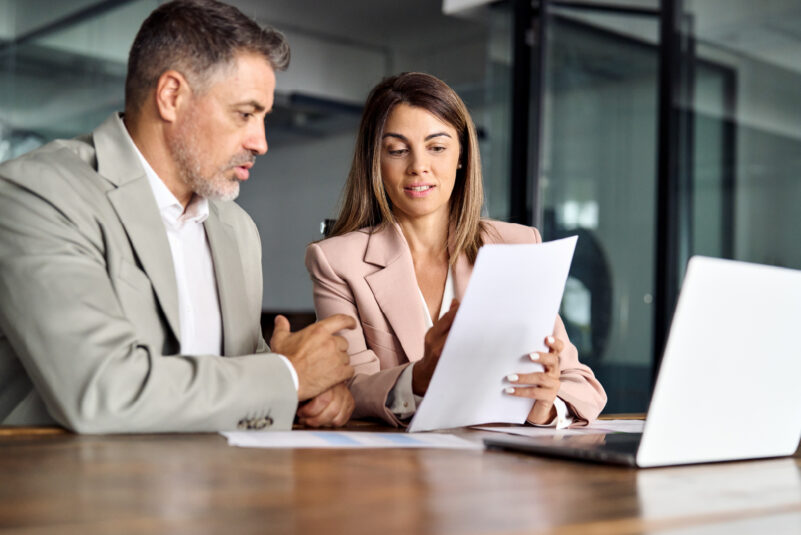 Blog_Are You Overpaying for Business Insurance- How an Audit Could Save You Thousands Two professionals sit at a table, discussing documents. A laptop is open in front of them with an office setting in the background.