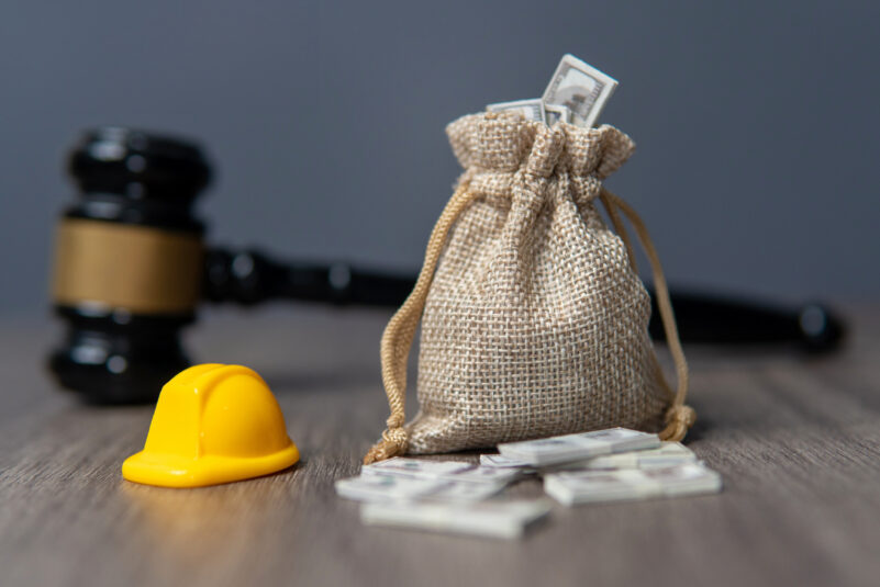 Blog_Workers’ Compensation Audits- The Mistake That Could Be Costing Your Business A gavel lies on a wooden surface behind a small burlap sack filled with money and a miniature yellow hard hat, symbolizing legal and financial matters in construction or labor.