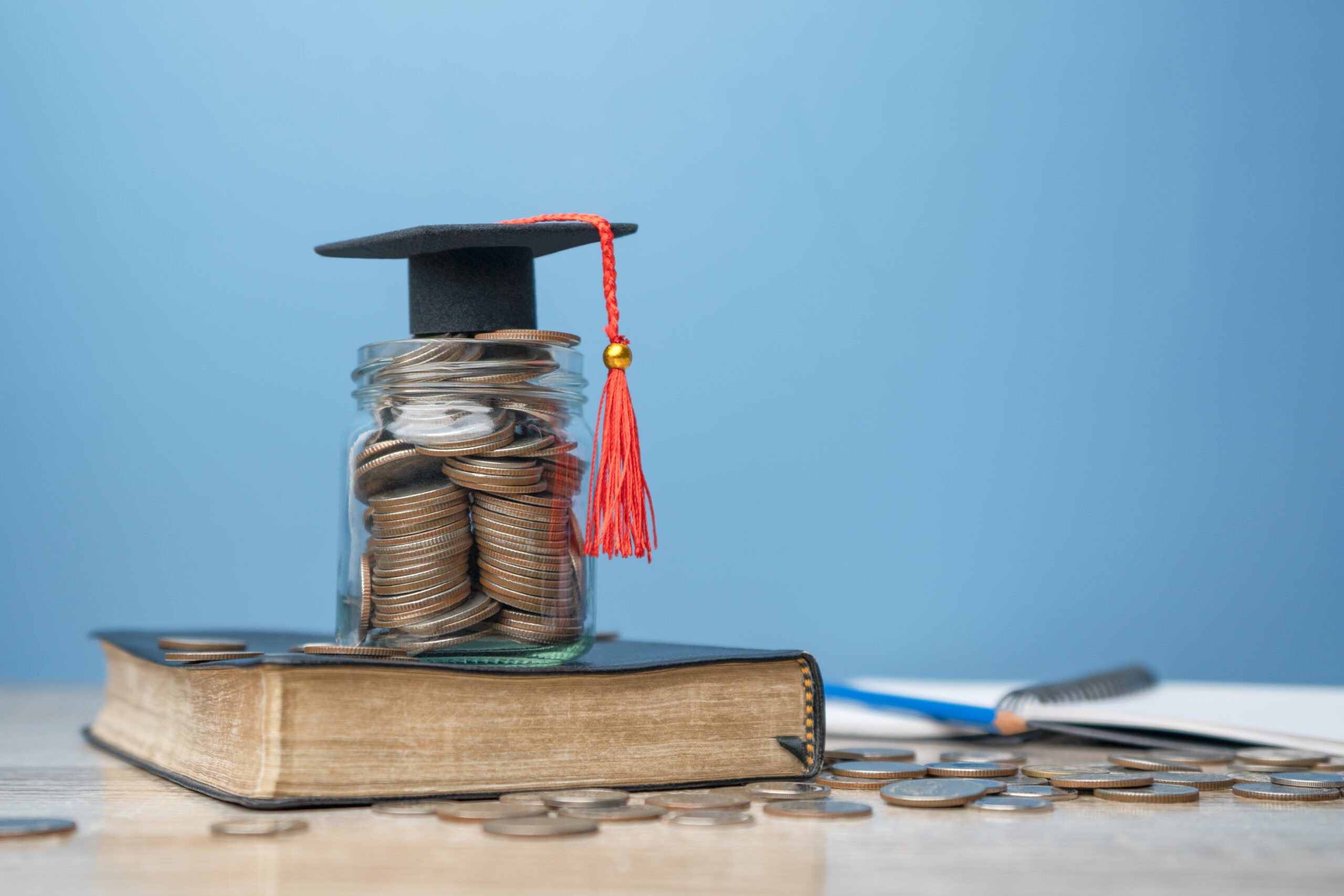 Saving,For,Student,Success,Scholarships,For,College,And,University,Tuition, A jar overflowing with coins, symbolizing savings for educational service, is elegantly topped with a graduation cap and rests on a book. Coins are scattered across the wooden surface, while a blue wall and an open notebook with a pen lie in the background.