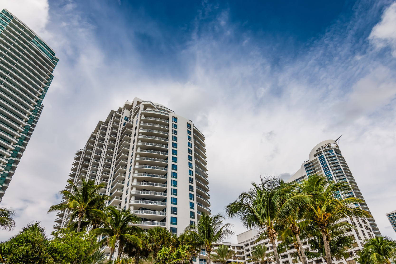Comegys-Blog_Navigating the Storm- 2025 Commercial Property Insurance Trends for Florida Business Owners Tall modern apartment buildings, reminiscent of a successful business funding venture, are surrounded by palm trees under a partly cloudy blue sky.
