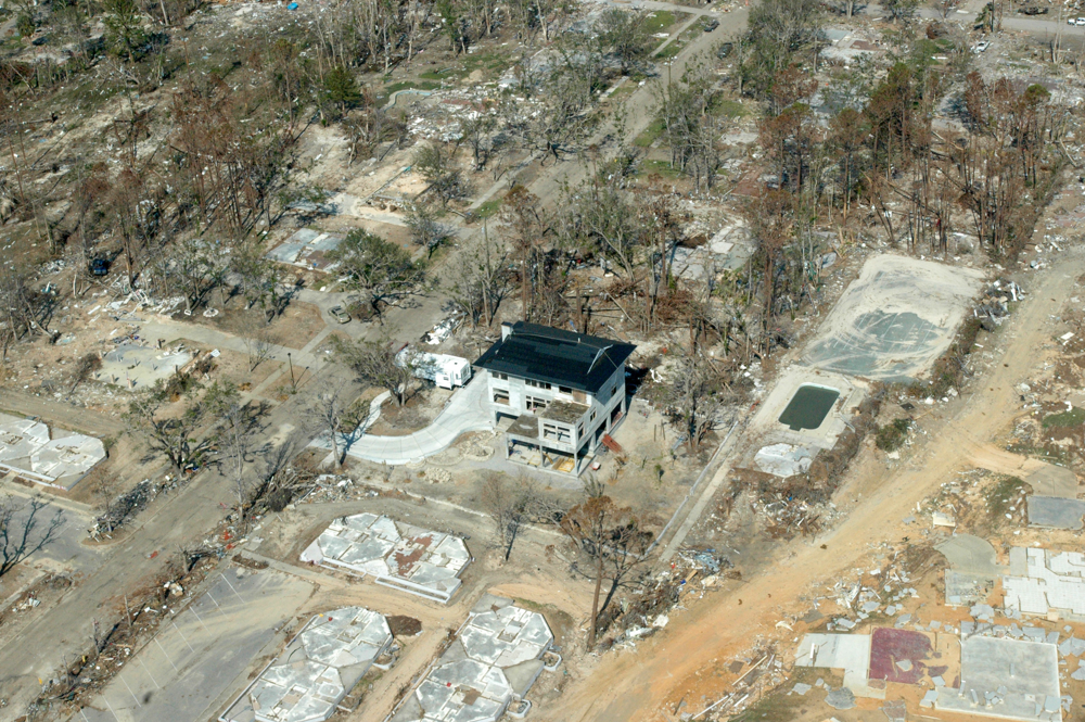 Aerial view of a mostly destroyed neighborhood with one intact house surrounded by debris, foundation slabs, scattered trees, and remnants of local businesses.