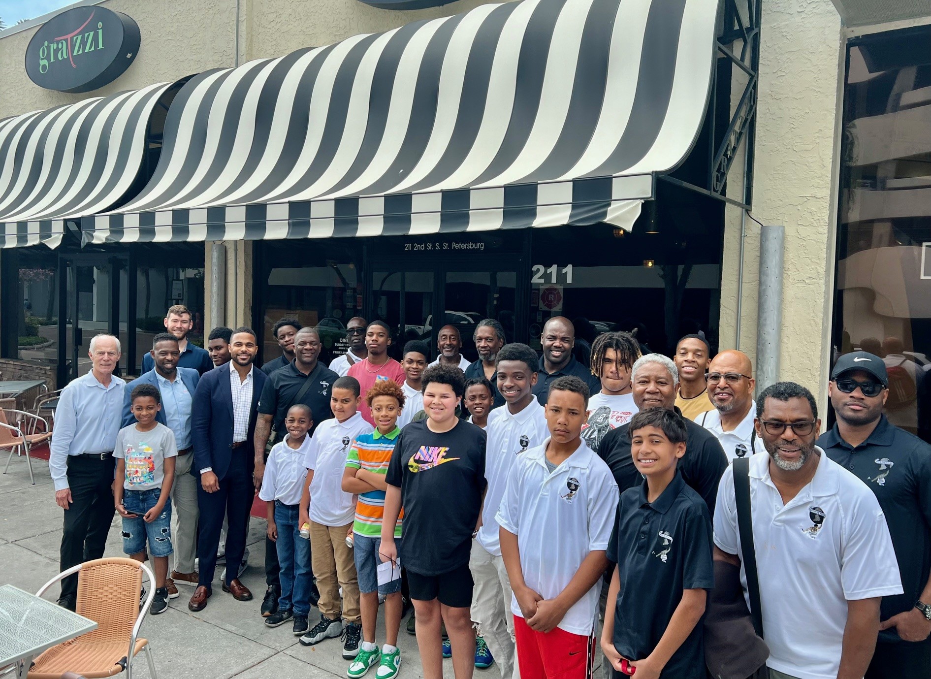 A diverse group of men and boys, including members of Men in the Making, pose for a photo outside a restaurant with a black-and-white striped awning and the sign "grazzi" above the entrance.