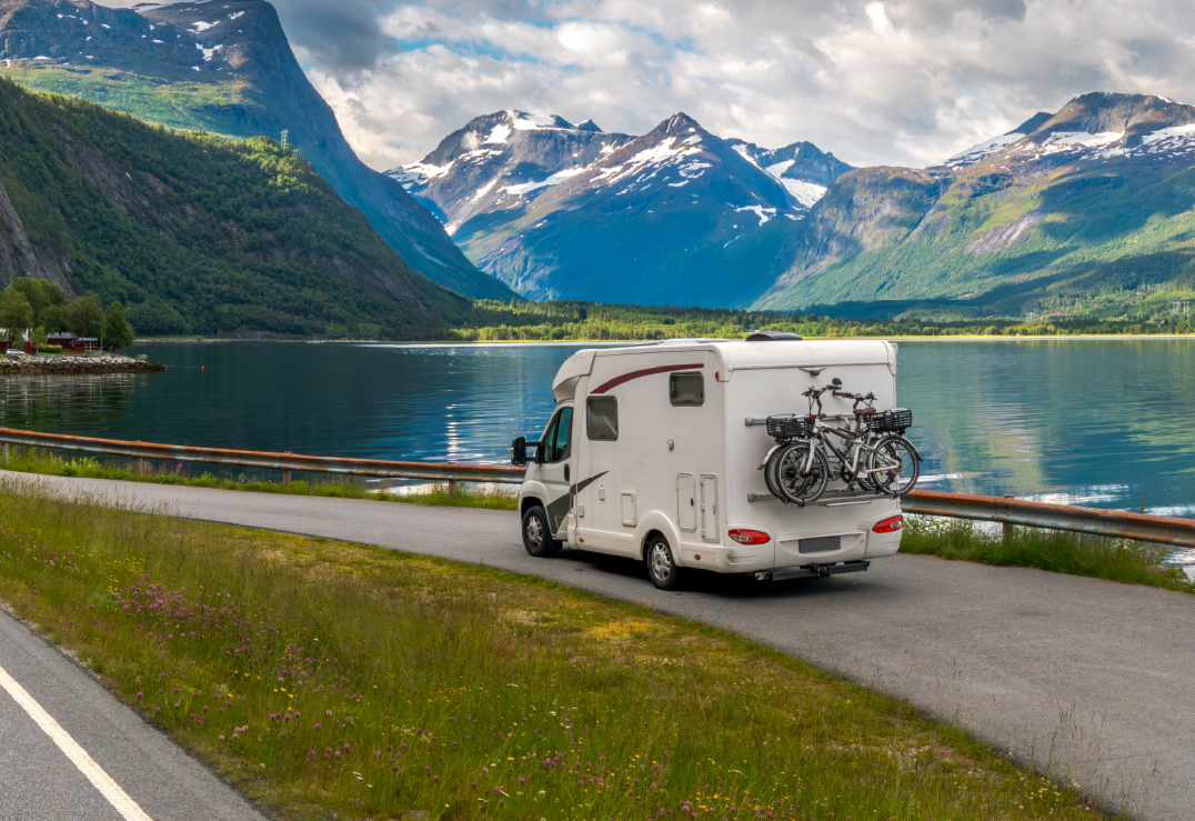 A white camper van with bicycles mounted on the back drives along a lakeside road, showcasing the freedom of motorhomes and travel, surrounded by mountains and greenery under a partly cloudy sky.