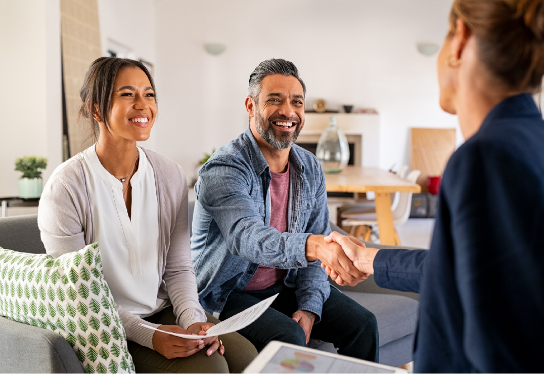 A man smiles and shakes hands with a woman across a table, while another woman sits beside him holding papers—everyone appears satisfied after choosing insurance at the agency.