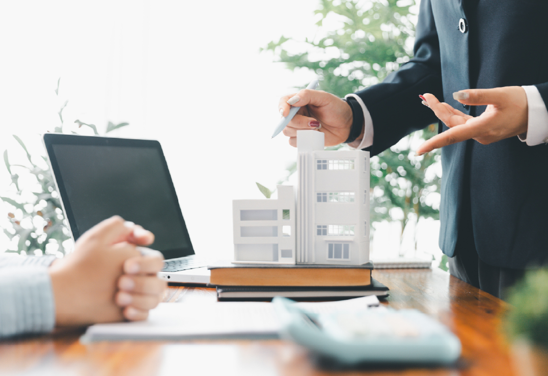 Two people discuss a building model on a desk with a laptop, documents, and plants in the background, exploring commercial insurance trends shaping the industry.