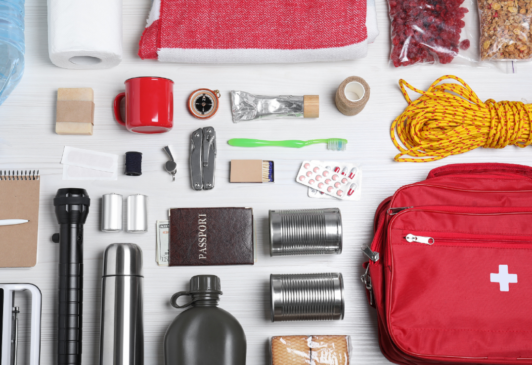 Essential emergency kit with assorted supplies for the family, including a red first aid kit, canned food, flashlight, rope, utensils, toiletries, water bottle, passport, and blanket arranged on a white surface.
