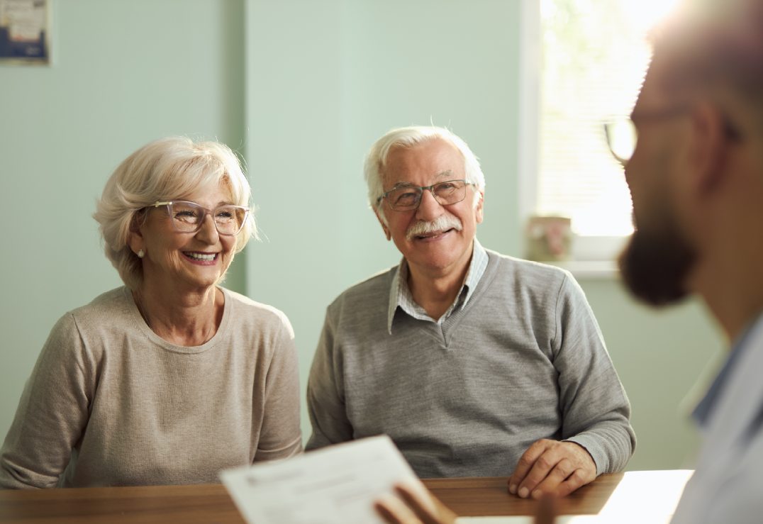 An elderly couple sits across a desk from a person holding medicare documents, having a conversation in a brightly lit room.