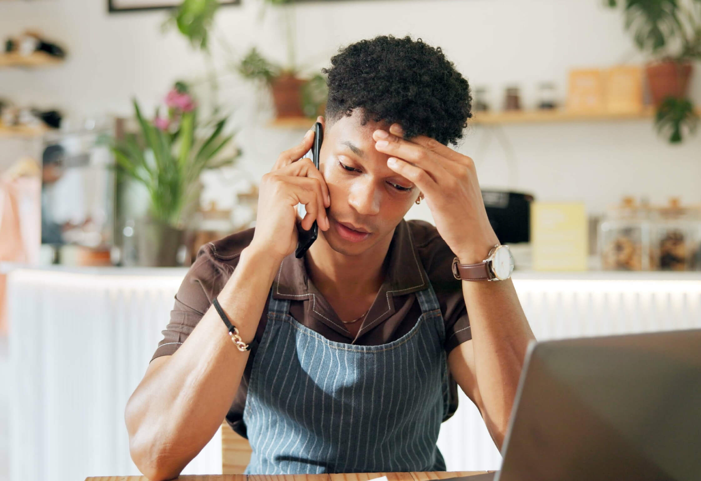A person wearing an apron talks on the phone while looking stressed, sitting at a table with a laptop in a cafe or kitchen setting—perhaps discussing SBA Loans, a common concern for Florida Business Owners and represents Hazard Insurance for SBA Loans: