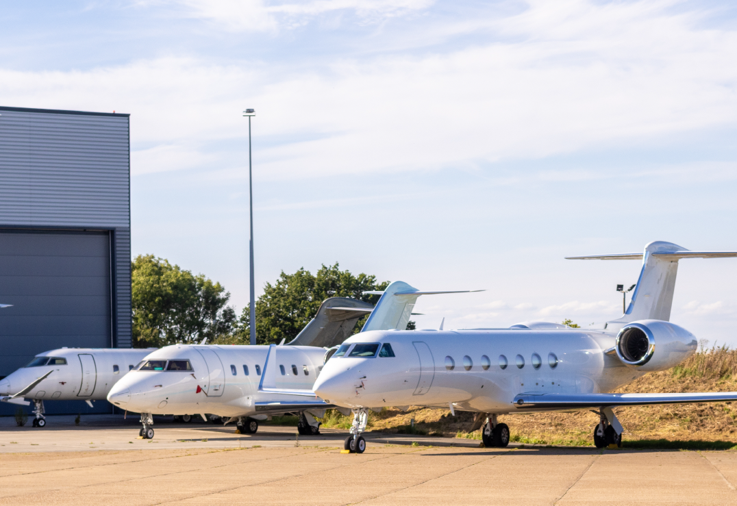 Three private jets are parked on a tarmac beside a hangar on a clear, sunny day, illustrating the reassurance aircraft insurance can provide for plane owners amid blue skies and trees in the background.