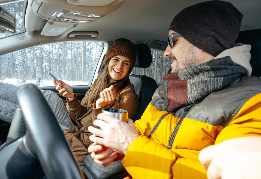 Two people sit in a car dressed in winter clothes; one holds a phone and smiles while the other, ready for holiday travel, wears sunglasses and holds a metal travel mug. Snowy trees are visible outside. Image represents holiday travel and auto insurance in Florida