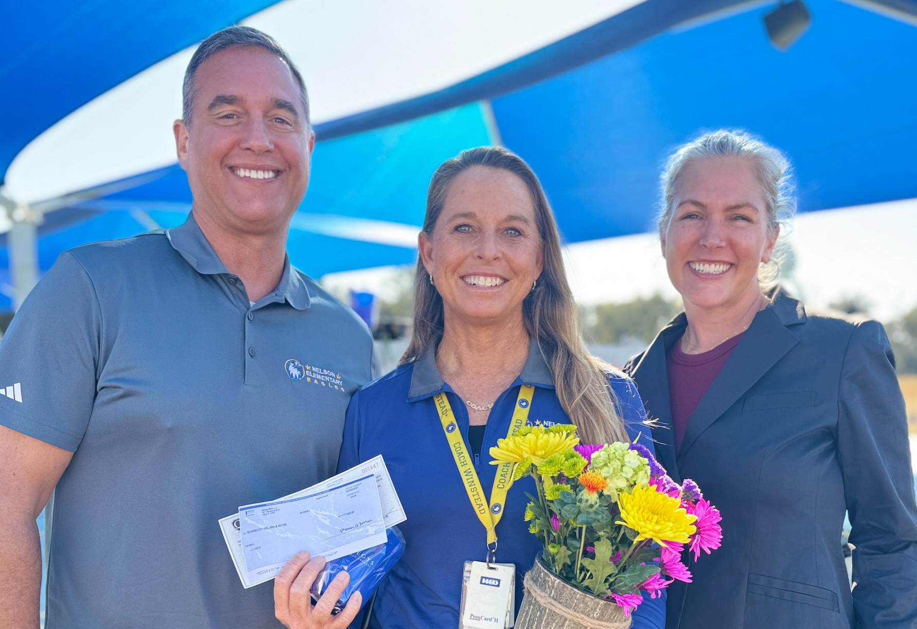Three adults stand together outdoors. The woman in the center, honored at Nelson Elementary School, holds a bouquet of flowers and papers, while the two beside her smile at the camera under a blue canopy.