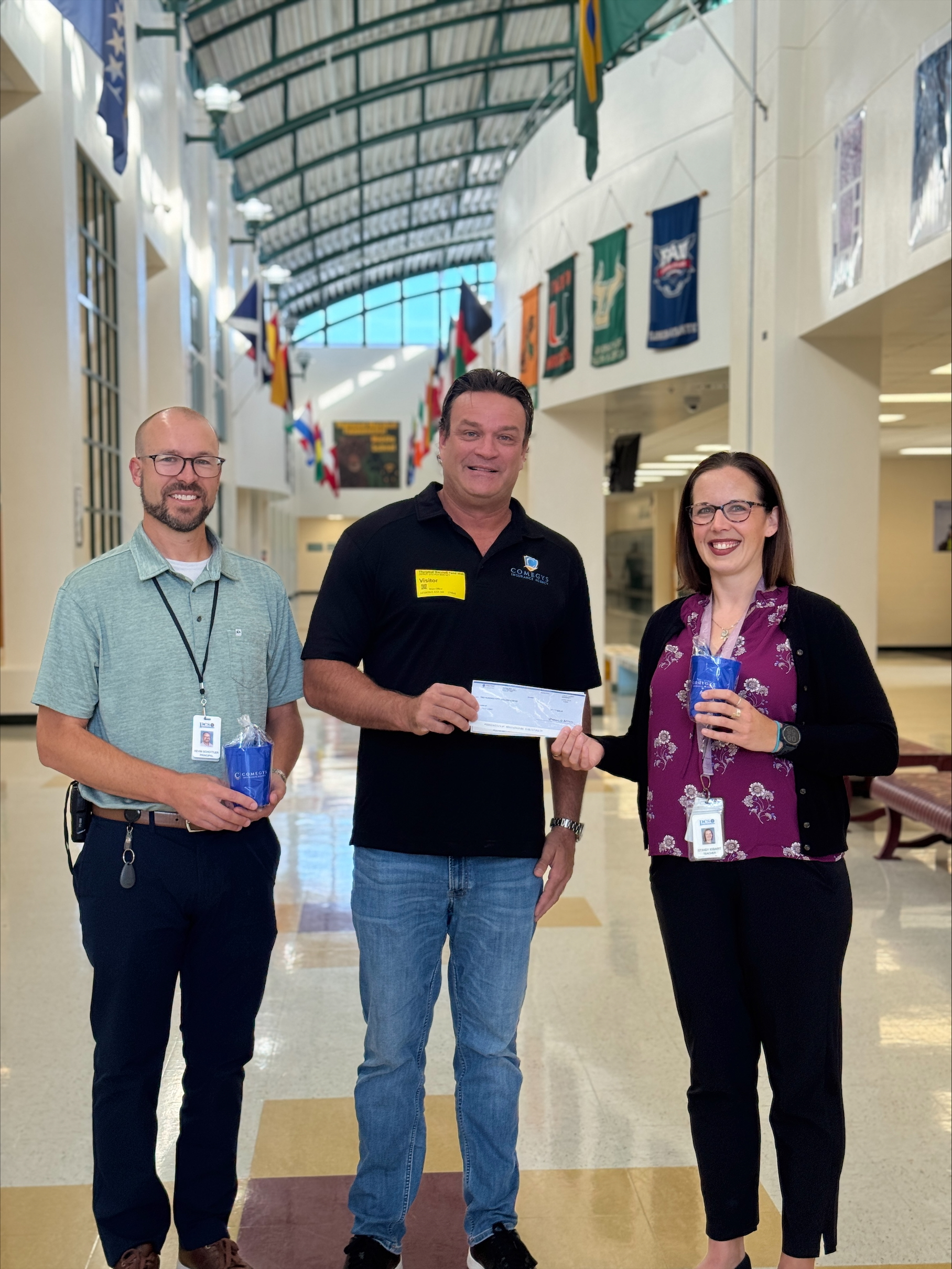 Three adults stand in a school hallway. The person in the middle holds a check, while the others hold blue tumblers from a Teacher Giveaways event. Flags hang from the ceiling above them.